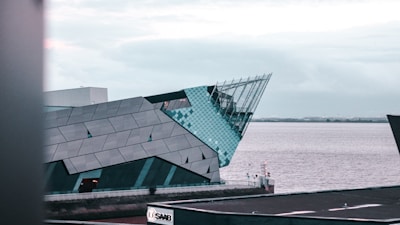 A modern architectural building with sharp angles, featuring a combination of metal and glass elements. The structure is situated near a body of water, with a cloudy sky overhead. In the foreground, there is a visible advertisement sign for Saab.