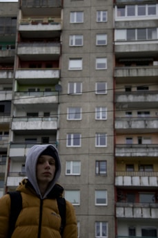 A person in a hooded jacket stands in front of a tall, multi-story apartment building with balconies. The building's exterior appears aged and weathered.