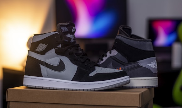 A pair of high-top sneakers is placed on a cardboard box. The sneakers feature a black, grey, and white color scheme with a distinct logo on the side. In the background, there is a blurred gradient of colors emanating from a monitor, adding a modern and sleek vibe.