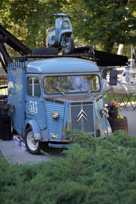 A vintage blue van is parked outdoors with a matching retro scooter mounted on its roof. The van's unique design features rounded headlights and a front grill emblem. A potted plant with colorful flowers and a basket of apples are placed beside the van. The background includes lush green trees and some outdoor furniture.