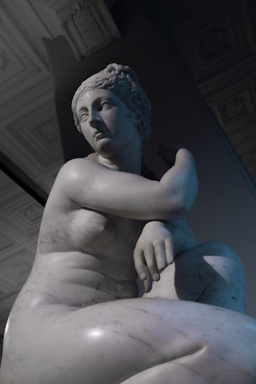 A marble sculpture of a contemplative figure with an intricate hairstyle and a serene expression. The figure is posed in a seated position with one arm resting on its knee, creating a relaxed yet elegant silhouette. The fine detailing in the facial features and the realistic texture of the marble highlight the craftsmanship.