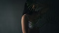 A woman in a white lace bra is partially obscured by a large palm leaf, creating a mysterious and intimate ambiance. The lighting is soft and dim, highlighting the contours of her arm and shoulder.