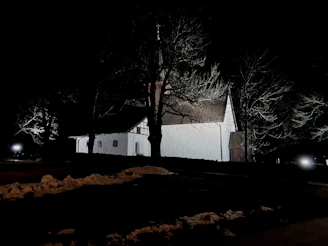 A white church with a pitched roof is illuminated at night, surrounded by barren trees. Sparse patches of snow are visible on the ground. The setting is dark, with minimal light highlighting the structure and the path leading to it.