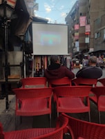 An outdoor urban setting with rows of red chairs facing a large screen displaying a projection. Two people are seated, watching the screen. Surrounding buildings are adorned with signs and banners. The area appears to be a street market or open space, with various stalls visible.