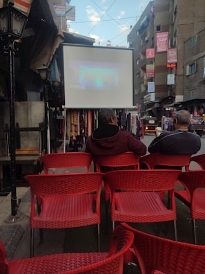 An outdoor urban setting with rows of red chairs facing a large screen displaying a projection. Two people are seated, watching the screen. Surrounding buildings are adorned with signs and banners. The area appears to be a street market or open space, with various stalls visible.