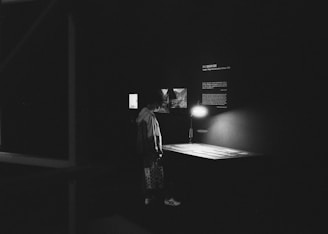 A dimly lit exhibition space shows a person standing in front of a small display table illuminated by a lamp. The area is surrounded by dark walls with a few lit-up photos or pieces of text present. The setting appears quiet and contemplative.