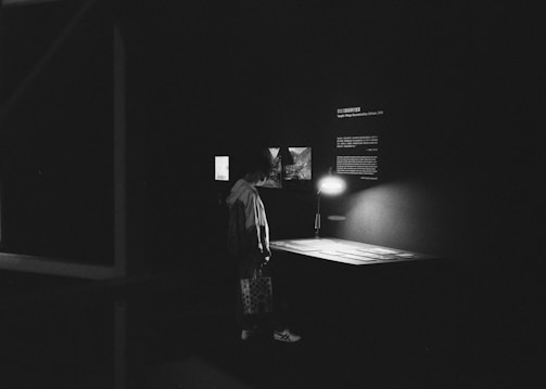 A dimly lit exhibition space shows a person standing in front of a small display table illuminated by a lamp. The area is surrounded by dark walls with a few lit-up photos or pieces of text present. The setting appears quiet and contemplative.