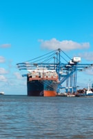 A large cargo ship is docked at a port with several large blue cranes extending over it. The ship is heavily loaded with stacked containers of various colors. The scene is set against a clear blue sky with a few scattered clouds, and the water in the foreground is calm.