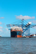 A busy port with cargo containers being carefully loaded onto a ship under a clear sky.