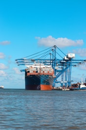 A large cargo ship is docked at a port with several large blue cranes extending over it. The ship is heavily loaded with stacked containers of various colors. The scene is set against a clear blue sky with a few scattered clouds, and the water in the foreground is calm.