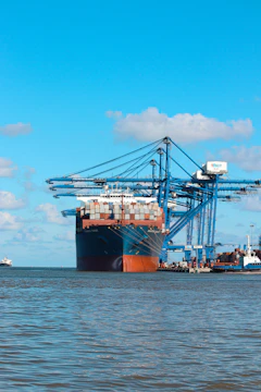 A cargo ship docked at a busy port with containers being loaded under a clear blue sky.