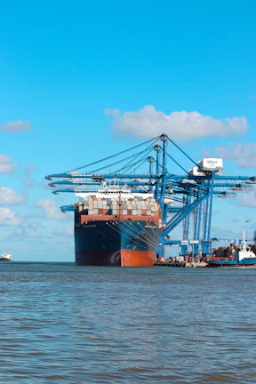 A large cargo ship docked at Al Khums port with containers being loaded by cranes under a navy blue sky.
