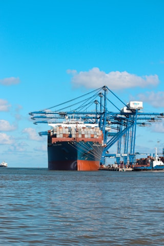 A large cargo ship is docked at a port with several large blue cranes extending over it. The ship is heavily loaded with stacked containers of various colors. The scene is set against a clear blue sky with a few scattered clouds, and the water in the foreground is calm.