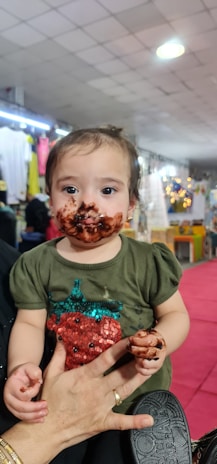 A small child with chocolate smeared on their face is being held by an adult. The child is wearing a green shirt with a colorful sequin strawberry design. The background appears to be indoors, with blurred lighting and decor, suggesting a lively or festive setting.