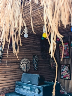 A rustic interior space with bamboo walls features tribal masks hanging in a row. There is a stereo system with an Onkyo speaker placed below the masks. Straw-like materials hang from the ceiling, and two light bulbs, one white and one yellow, are visible. A colorful movie poster with Hindi text decorates the wall.