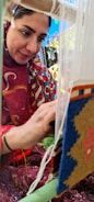 A woman intently weaving a colorful tapestry on a loom. She is adorned in a patterned headscarf and traditional attire, concentrating on her craft amidst vibrant colors and fabric patterns.