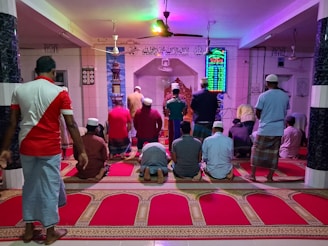 A group of people are engaged in prayer inside a mosque. They are kneeling on ornate red prayer rugs and facing towards an intricately decorated niche. The walls are adorned with decorative text and a digital clock displaying prayer times. Some individuals wear traditional attire and caps.