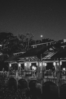 A nighttime scene featuring a wooden structure or pavilion with ornate railings, illuminated by several lights. Trees and a crescent moon are visible in the background, creating a serene and picturesque atmosphere.