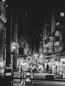A bustling city street at night, featuring numerous people walking and shopping in a lively market area. Bright signs illuminate the area, highlighting the vibrant atmosphere. Buildings flank the street, filled with various shops and stalls offering goods and services.