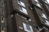 Close-up view of a tall, Brutalist-style concrete apartment building with rows of windows and balconies. The architecture features prominent use of raw concrete and repetitive patterns. Several windows appear to have ventilation grills or air conditioning units.