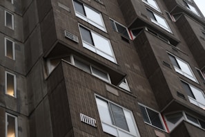 Close-up view of a tall, Brutalist-style concrete apartment building with rows of windows and balconies. The architecture features prominent use of raw concrete and repetitive patterns. Several windows appear to have ventilation grills or air conditioning units.