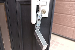 A metallic lockbox is attached to the handle of a black door. The front panel of the lockbox is open, revealing its interior with buttons and a mechanism inside. The door has a cylindrical lock above the handle, and the edge of a brown wall is visible on the right.