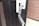 A metallic lockbox is attached to the handle of a black door. The front panel of the lockbox is open, revealing its interior with buttons and a mechanism inside. The door has a cylindrical lock above the handle, and the edge of a brown wall is visible on the right.