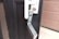 A metallic lockbox is attached to the handle of a black door. The front panel of the lockbox is open, revealing its interior with buttons and a mechanism inside. The door has a cylindrical lock above the handle, and the edge of a brown wall is visible on the right.