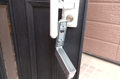 A metallic lockbox is attached to the handle of a black door. The front panel of the lockbox is open, revealing its interior with buttons and a mechanism inside. The door has a cylindrical lock above the handle, and the edge of a brown wall is visible on the right.