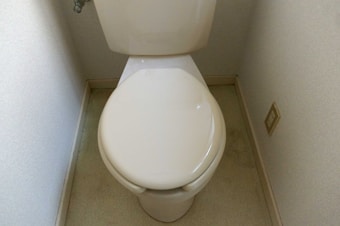A clean, white-tiled toilet in a small, narrow bathroom space. The walls are plain, and there's a power outlet nearby.