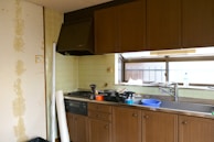 A kitchen space with wooden cabinets and a stainless steel sink. The countertop features various items including a blue plastic basin, a bottle, and some clutter. An exhaust hood is installed above the stove, which appears to have some cooking utensils. The walls are tiled, and a section shows peeling wallpaper. Through the window, a railing can be seen outside.