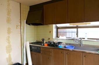 A kitchen space with wooden cabinets and a stainless steel sink. The countertop features various items including a blue plastic basin, a bottle, and some clutter. An exhaust hood is installed above the stove, which appears to have some cooking utensils. The walls are tiled, and a section shows peeling wallpaper. Through the window, a railing can be seen outside.