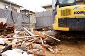 A construction site with a large amount of debris including wood and metal pieces scattered across the ground. A yellow excavator with visible treads is parked amidst the rubble. Surrounding the area are gray tarps and a residential building in the background.