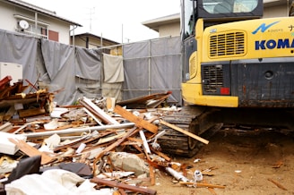 A construction site with a large amount of debris including wood and metal pieces scattered across the ground. A yellow excavator with visible treads is parked amidst the rubble. Surrounding the area are gray tarps and a residential building in the background.