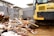 A construction site with a large amount of debris including wood and metal pieces scattered across the ground. A yellow excavator with visible treads is parked amidst the rubble. Surrounding the area are gray tarps and a residential building in the background.