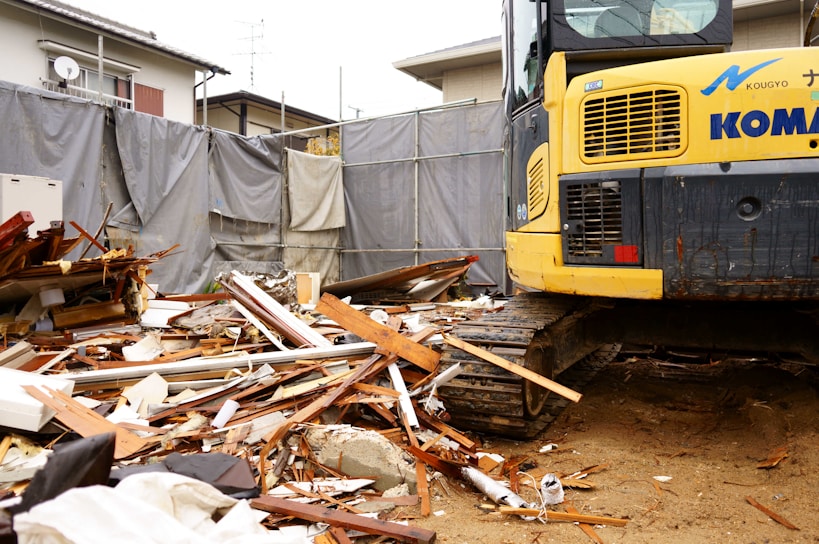 A construction site with a large amount of debris including wood and metal pieces scattered across the ground. A yellow excavator with visible treads is parked amidst the rubble. Surrounding the area are gray tarps and a residential building in the background.