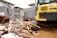 A construction site with a large amount of debris including wood and metal pieces scattered across the ground. A yellow excavator with visible treads is parked amidst the rubble. Surrounding the area are gray tarps and a residential building in the background.