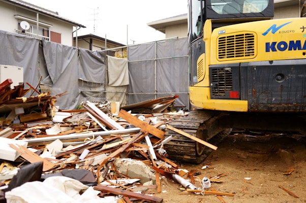 A construction site with a large amount of debris including wood and metal pieces scattered across the ground. A yellow excavator with visible treads is parked amidst the rubble. Surrounding the area are gray tarps and a residential building in the background.