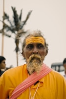 An elderly man with a large white beard and wearing an orange robe is seen with a yellow painted forehead. He carries a pink cloth diagonally across his body. The background features out-of-focus people and a palm tree.