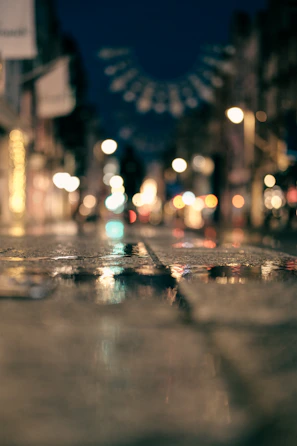 An urban street scene at dusk with colorful lights reflecting on wet pavement.