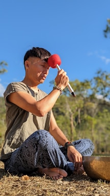 A person is seated on the ground in a meditative pose, holding a red-tipped percussion mallet to their forehead. They are wearing a beige shirt and loose blue patterned pants. In front of them, a large singing bowl is placed on the ground. The background features a clear blue sky and some green trees.