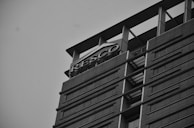 A tall, modern building with multiple floors and sleek architectural design is captured from a low angle. The structure features large windows arranged in horizontal lines, and there is a prominent sign with the word 'RESCO' at the top. The image is in grayscale, contributing to a slightly somber or formal atmosphere.