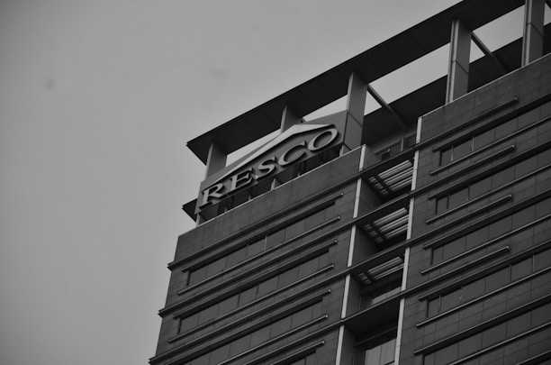 A tall, modern building with multiple floors and sleek architectural design is captured from a low angle. The structure features large windows arranged in horizontal lines, and there is a prominent sign with the word 'RESCO' at the top. The image is in grayscale, contributing to a slightly somber or formal atmosphere.