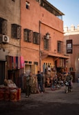 A street scene in a traditional market area featuring various textiles and leather goods displayed outside a shop. The architecture includes warm-toned walls with ornate window grilles. Two people are standing by the shop, and two others are on motorcycles. The ambiance suggests a typical day in a bustling marketplace.