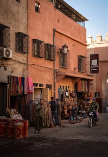 A street scene in a traditional market area featuring various textiles and leather goods displayed outside a shop. The architecture includes warm-toned walls with ornate window grilles. Two people are standing by the shop, and two others are on motorcycles. The ambiance suggests a typical day in a bustling marketplace.