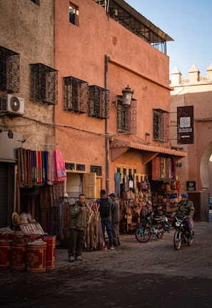 A street scene in a traditional market area featuring various textiles and leather goods displayed outside a shop. The architecture includes warm-toned walls with ornate window grilles. Two people are standing by the shop, and two others are on motorcycles. The ambiance suggests a typical day in a bustling marketplace.