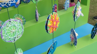 Colorful, handcrafted circular decorations hang on strings against a vivid green background. Each circle is intricately woven with yarn in various patterns and colors. The display is arranged on steps with alternating blue and green surfaces, creating a playful and artistic atmosphere.
