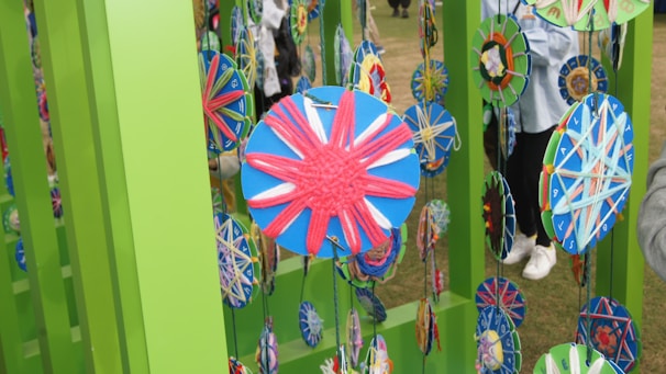Colorful circular crafts are suspended from vibrant green frames, featuring intricate patterns made with yarn in various colors. The background includes partially visible people and a grassy area.