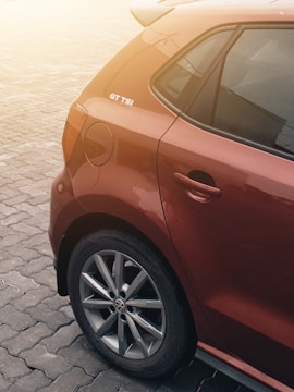A red car with a GT TSI badge is parked on a paved surface. The image highlights the rear side of the car, focusing on the wheel with a prominent logo and the detail in the paint. The lighting suggests a warm, slightly overcast environment.