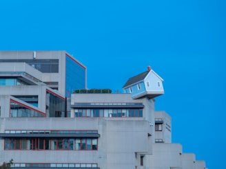 A unique architectural scene with a small white house perched precariously on the edge of a modern concrete building. The building has multiple levels with a minimalist design and red trim around the windows, set against a clear blue sky.